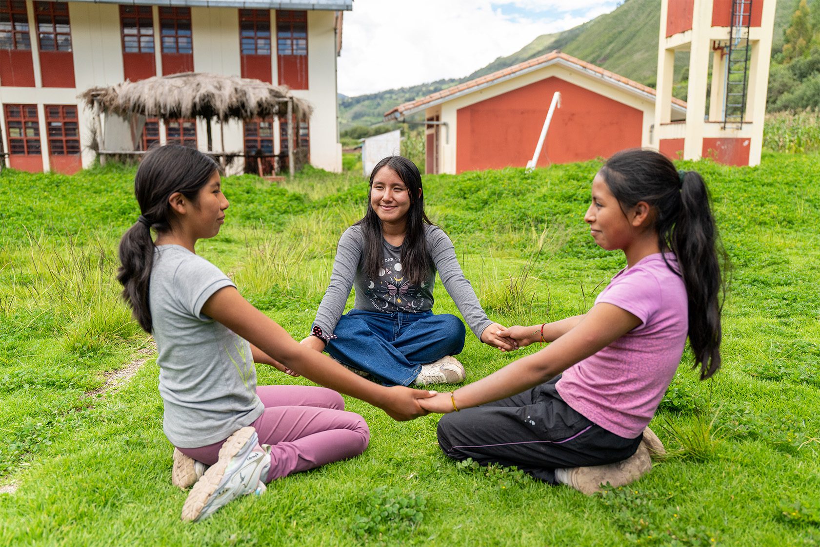 Three girls sit in circle on the grass, holding hands 