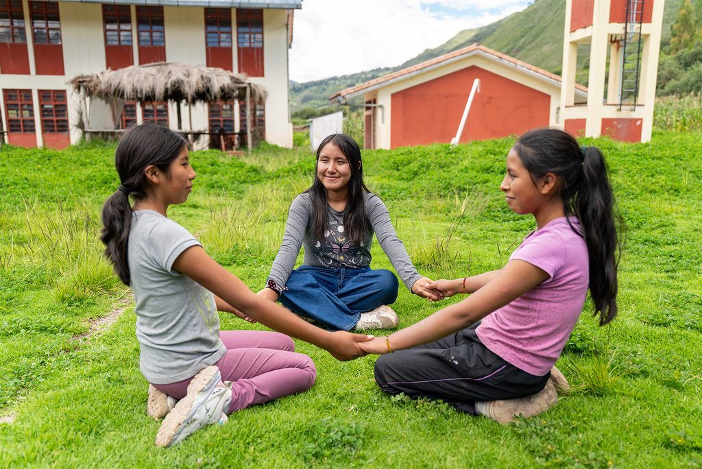 Three girls sit in circle on the grass, holding hands
